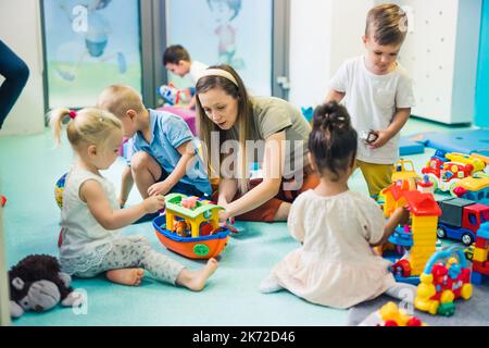 Insegnante caucasico biondo che aiuta i bambini a costruire le cose correttamente nel vivaio, un parco giochi pieno di giocattoli, attività creativa per i bambini. Foto di alta qualità Foto Stock