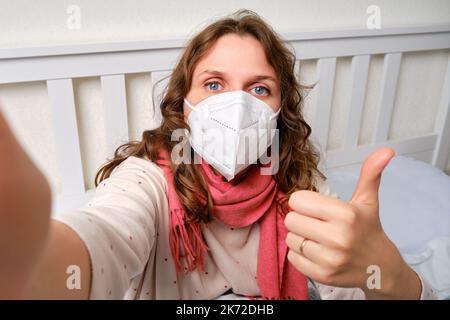 Donna malata blogger prende selfie foto sul telefono per il blog online. Donna ammalata adulta avvolta in una sciarpa rossa siede su un letto domestico in una camera da letto bianca, femmina A. Foto Stock