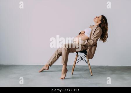 Donna incinta seduta sulla sedia in studio con sfondo bianco. Donna incinta che tocca il ventre Foto Stock