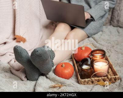 Una ragazza in un maglione e calzini a maglia si siede su una coperta e lavora su un computer portatile. Accanto a lei c'è un vassoio con tutte le cose e le zucche. Accogliente concetto autunnale. Foto Stock