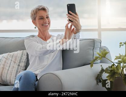 Vivere la sua vita migliore. una donna anziana attraente che prende selfie mentre si rilassa nel suo salotto a casa. Foto Stock
