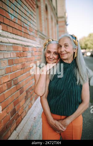 Donne anziane gemelle, negli stessi vestiti in piedi e posando di fronte al muro di mattoni, all'aperto in città. Foto Stock