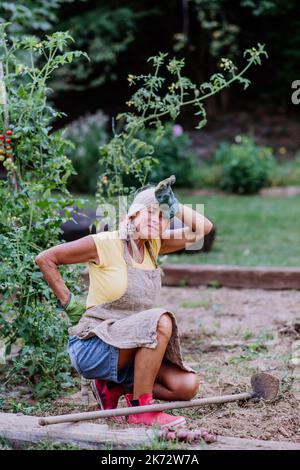 Donna anziana che riposa dopo aver lavorato manualmente nel suo giardino, avendo dolore alla schiena. Foto Stock