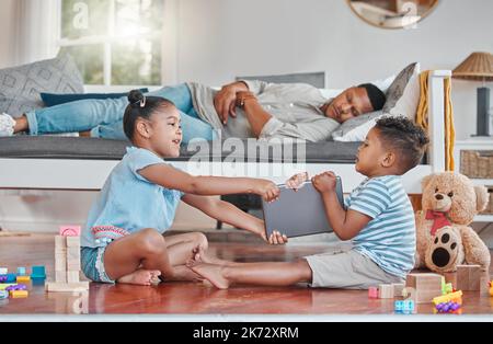 Papà dice il suo turno. due fratelli combattono su un tablet digitale sul pavimento mentre il loro papà dorme sul divano a casa. Foto Stock