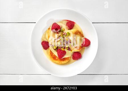 Frittelle dolci al formaggio con crema di cocco, lamponi e pistacchi su superficie di legno bianco Foto Stock