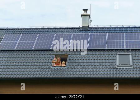 Madre felice e suo figlio che ondeggiavano dalla finestra del lucernario nella loro nuova casa con pannelli solari sul tetto. Energia alternativa, risparmio di risorse e. Foto Stock