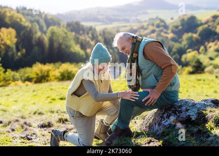 Uomo anziano con ginocchio ferito riposarsi durante la passeggiata autunnale, sua moglie si prende cura di lui. Foto Stock