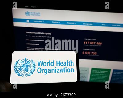 Persona che tiene il cellulare con il logo dell'agenzia ONU Organizzazione Mondiale della Sanità (OMS) sullo schermo di fronte alla pagina web. Messa a fuoco sul display del telefono. Foto Stock