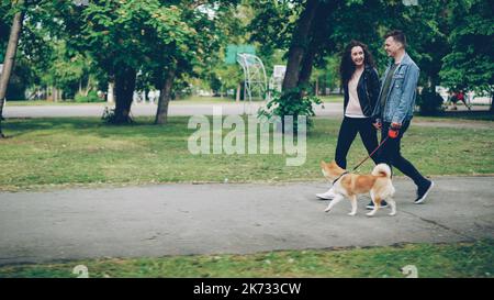 Allegro giovane coppia uomo e donna stanno camminando il cane nel parco vicino a campi sportivi, la gente sta tenendo le mani, il ragazzo è alla guida dell'animale. Vita urbana, relazione e concetto di animali domestici. Foto Stock