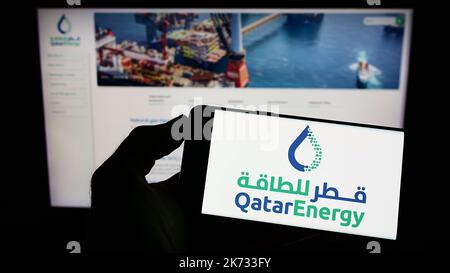 Persona che tiene il cellulare con il logo della società petrolifera Qatar QatarEnergy sullo schermo di fronte al sito web aziendale. Messa a fuoco sul display del telefono. Foto Stock