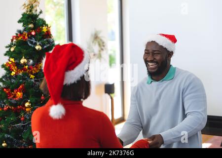 Felice coppia diversa indossando cappelli di babbo natale, ballando accanto all'albero di natale Foto Stock