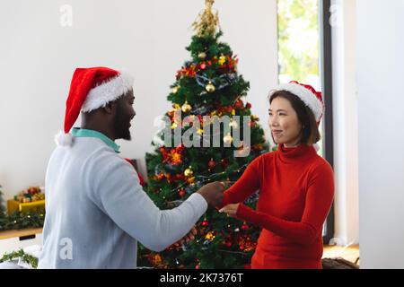 Felice coppia diversa indossando cappelli di babbo natale, ballando accanto all'albero di natale Foto Stock