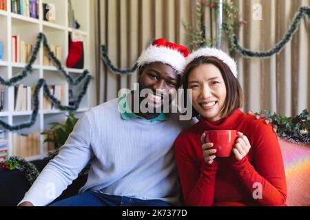 Ritratto di felice coppia diversa indossando cappelli di babbo natale guardando la macchina fotografica e sorridendo Foto Stock