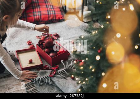 Bambina addobba l'abete di Natale con palline ornamentali rosse. Piccolo capretto che tiene il regalo in mano prima di appenderlo all'albero con la luce, garland. Scatola con palline appoggiate sul pavimento con decorazioni Foto Stock