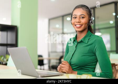 Headshot di giovane dipendente afroamericano che indossa cuffie wireless, guardando la fotocamera con un sorriso. Donna allegra in cuffie con microfono, supporto tecnico in ufficio Foto Stock