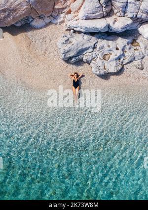 Foto vista dall'alto che mostra un costume da bagno nero vestito da donna sdraiato su una spiaggia di ciottoli bianchi solitari, onde calme che le lavano le gambe. Vacanza esotica estiva tropicale Foto Stock