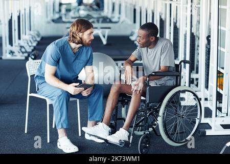 Giovane medico in uniforme che discute la riabilitazione con il paziente mentre si siede in sedia a rotelle in palestra Foto Stock