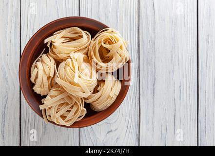 Tagliatelle non cotte in ciotola di ceramica su sfondo bianco in legno Foto Stock