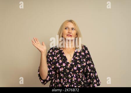 Bella donna in abito a motivi scuri in piedi su sfondo beige sorridendo gentilmente offrendo qualcosa con mano aperta o stretta di mano come saluto Foto Stock