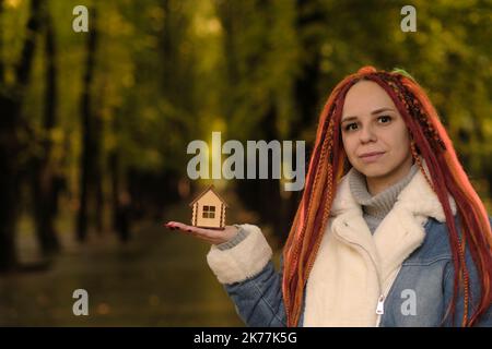 Donna positiva con greggi che tiene piccola casa in legno nel parco forestale. Donna con la casa dei giocattoli che guarda la macchina fotografica, sorridente. Concetto di acquisto nuovo Foto Stock