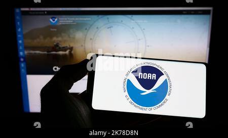 Persona che tiene il cellulare con il logo della National Oceanic and Atmospheric Administration (NOAA) sullo schermo di fronte alla pagina web. Messa a fuoco sul display del telefono. Foto Stock