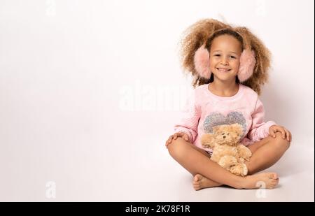 bellissimo bambino afroamericano che indossa cuffie, cappello che abbraccia orsetto di orsacchiotto e sorridente alla macchina fotografica isolata su bianco Foto Stock