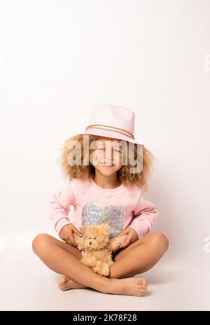 bellissimo bambino afroamericano che indossa un cappello di paglia che abbraccia un orsetto di peluche e sorride alla macchina fotografica isolata su bianco Foto Stock