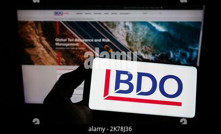 Persona che tiene il telefono cellulare con il logo della rete di contabilità BDO Global sullo schermo di fronte alla pagina web aziendale. Messa a fuoco sul display del telefono. Foto Stock