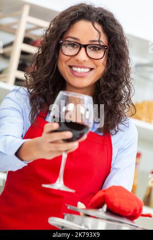 Bella ragazza o ragazza Hispanic felice Latina con denti perfetti indossando un grembiule rosso bere un bicchiere di vino rosso e cucinare nella sua cucina a. Foto Stock