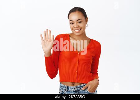 Ragazza nera amichevole, sorridente e salutando, agitando la mano a voi, benvenuto, in piedi su sfondo bianco Foto Stock