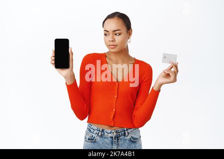 Donna delusa che mostra la carta di credito, guardando sconvolto l'app per lo schermo del telefono cellulare, accigliato, in piedi su sfondo bianco Foto Stock