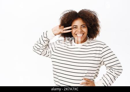 Sorridente ragazza ameirca africana positiva mostra la pace, segno di V vicino agli occhi e sembra felice, si trova su sfondo bianco Foto Stock