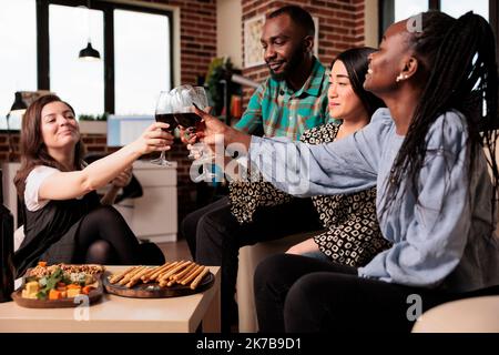 Etnie diverse gruppo di amici, brinda, bere vino, mangiare spuntini, divertirsi al compleanno di un giovane adulto nel soggiorno festa appartamento. Persone multiculturali che godono di amicizia. Foto Stock