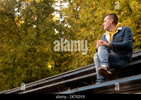Giovane uomo positivo con greggi che si guarda via, sorridente, seduto su vecchia panchina di strada bleacher in autunno dorato Foto Stock