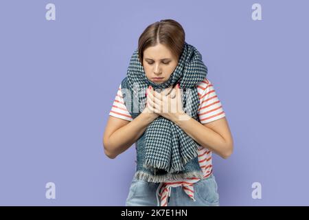 Ritratto di donna bionda malata malsana che indossa una T-shirt a righe e avvolta in sciarpa che soffre mal di gola, tosse, femmina che cattura freddo. Studio al coperto isolato su sfondo viola. Foto Stock