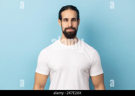 Ritratto dell'uomo comico positivo con barba che indossa una T-shirt bianca con un occhio a croce, divertendosi con l'espressione sciocca del volto, giocando a pazzo. Studio in interni isolato su sfondo blu. Foto Stock