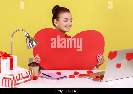 Donna felice che tiene un grande cuore di carta rossa e sorridente, seduto sul posto di lavoro con il laptop in cuori appiccicosi, videochiamata. Studio interno girato isolato su sfondo giallo. Foto Stock