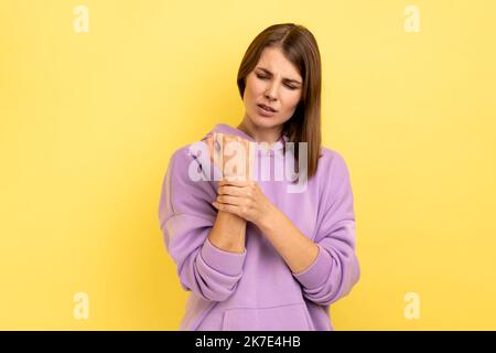 Ritratto di donna sconvolta in piedi con dolore, massaggiare il polso irritato, subire lesioni alle mani o spruce, indossare felpa con cappuccio viola. Studio al coperto isolato su sfondo giallo. Foto Stock