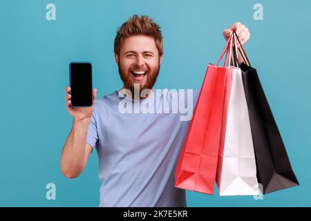 Ritratto di uomo ottimista bearded in piedi con lo sguardo soddisfatto che mostra le borse di shopping e smartphone con schermo vuoto per la pubblicità. Studio in interni isolato su sfondo blu. Foto Stock