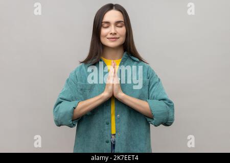 Donna splendida tiene le mani in un gesto di yoga, ha espressione facciale calma, mantenendo le palme pressate insieme e gli occhi chiusi, indossando giacca casual stile. Studio in interni isolato su sfondo grigio. Foto Stock