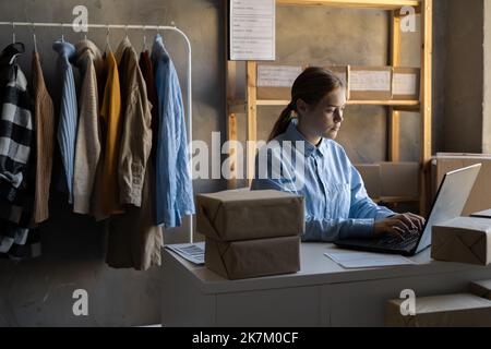 Venditore con computer portatile e pacchi in ufficio. Negozio online, shopping di abbigliamento di moda spedito in scatole di cartone di spedizione. Foto Stock