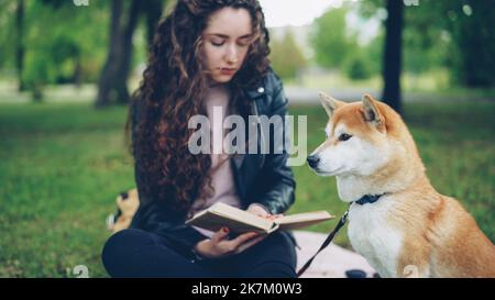 Bella giovane signora sta leggendo il libro nel parco e pattugliando carino cane shiba inu razza, ben allevato animale domestico è seduto sul prato vicino al suo proprietario. Concetto di animali, natura e persone. Foto Stock