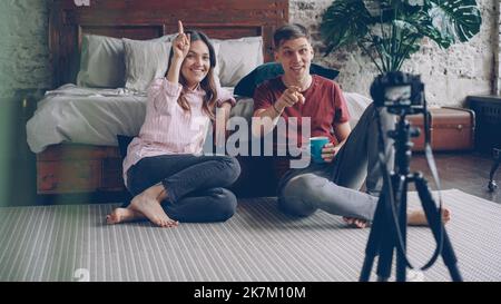 Attraente giovane marito e moglie blogger stanno registrando video vlog, agitando le mani e gesturing, parlando e ridendo, stanno usando la macchina fotografica su treppiede e seduto sul pavimento della camera da letto. Foto Stock