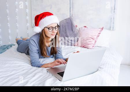 Bella donna seduta a letto che lavora con un computer portatile indossando il cappello di babbo natale per Natale con un sorriso felice e fresco sul viso. Chatta tramite videochiamata o acquista regali durante il Black Friday per Natale Foto Stock