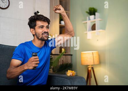 Un giovane entusiasta che urla alzando le mani mentre guarda una partita di sport in diretta in tv o in televisione a casa - concetto di intrattenimento, relax e. Foto Stock