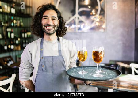 Amichevole ritratto di giovane cameriere sicuro maschio che tiene vassoio di servizio con birra Foto Stock