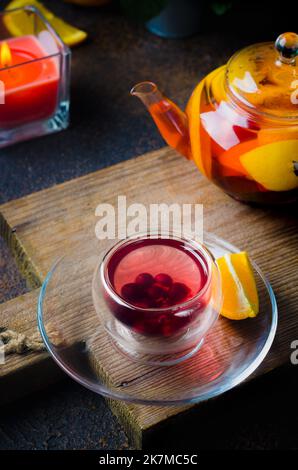 Tè alla frutta aromatizzato in casa in una tazza di vetro e teiera con arancia e limone appena sfornati, bacche, menta e Miele su sfondo rustico scuro. Caldo Foto Stock