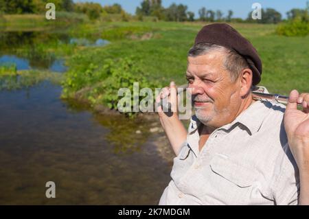 Ritratto di poaitive ucraino anziano uomo seduto su un fiume e sorridente mentre prende bastone camminare sul collo Foto Stock
