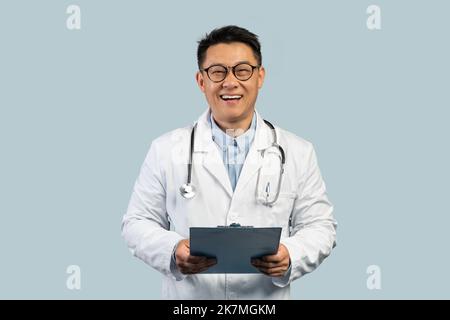Allegro uomo coreano di mezza età in cappotto bianco e bicchieri con tavoletta, isolato su sfondo blu Foto Stock