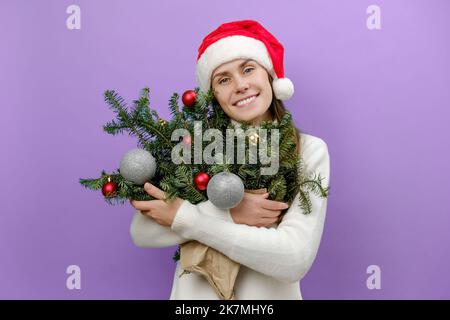 Ritratto di bella giovane donna felice indossando maglione bianco accogliente e cappello di Natale con abbraccio bouquet di rami di abete rosso, isolato su sfondo viola Foto Stock